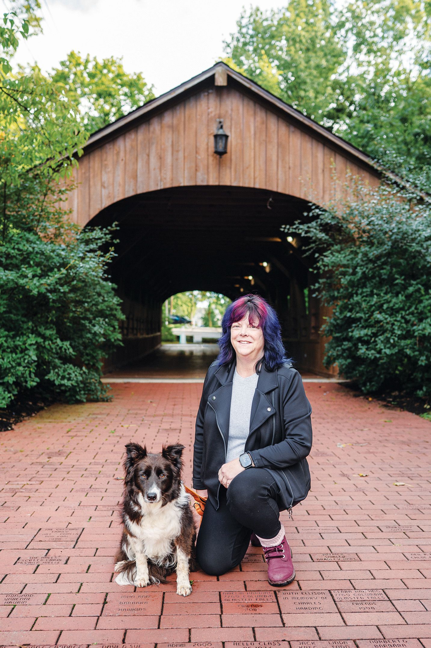 A person with purple-streaked hair kneels on a brick path with a dog, posing in front of a wooden covered bridge.