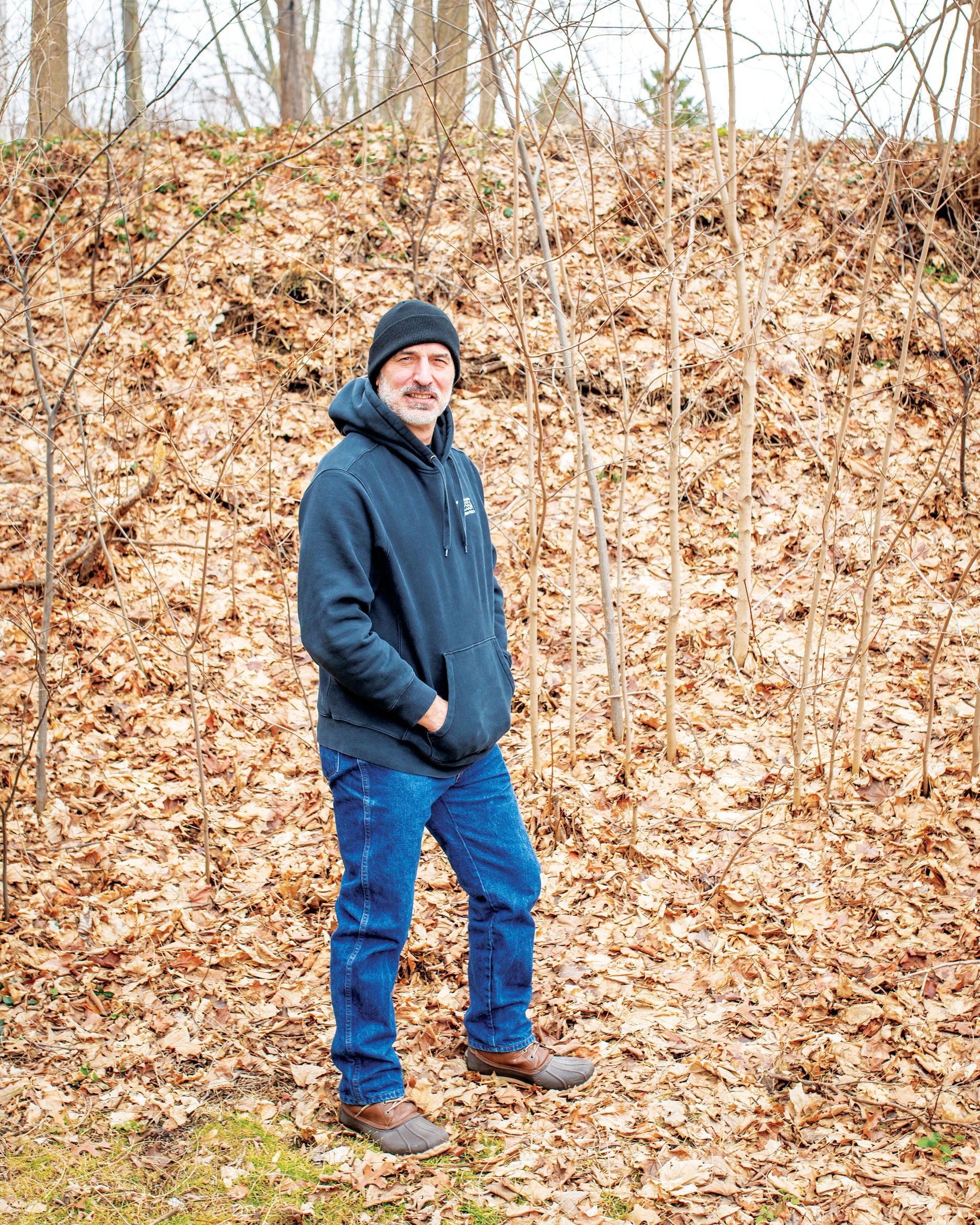 A person with a mustache wearing a black hoodie, beanie, and jeans standing on a leaf-covered slope in a wooded area.