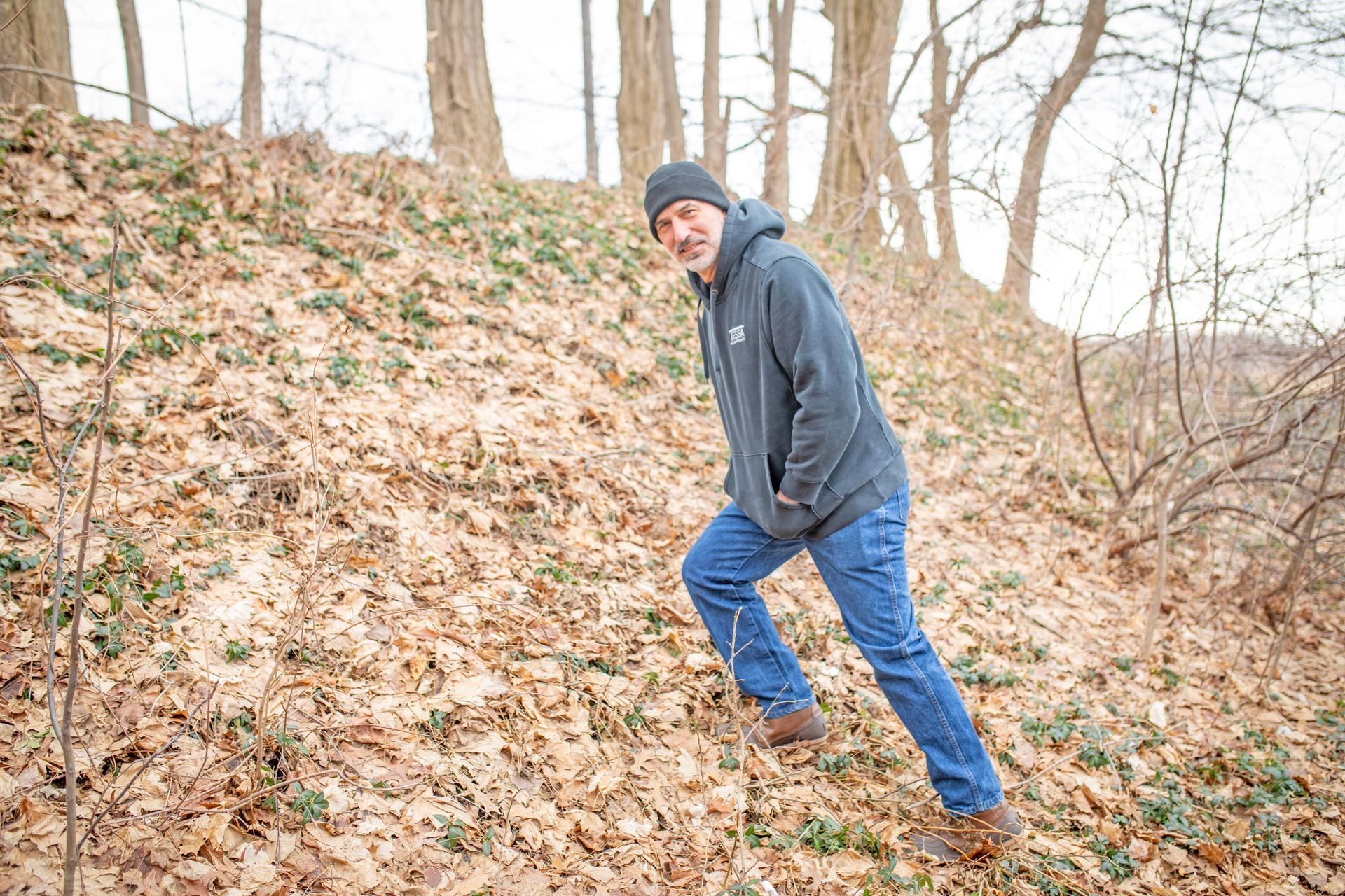 A person in a gray hoodie and jeans walks up a steep, leaf-covered embankment in a wooded area.