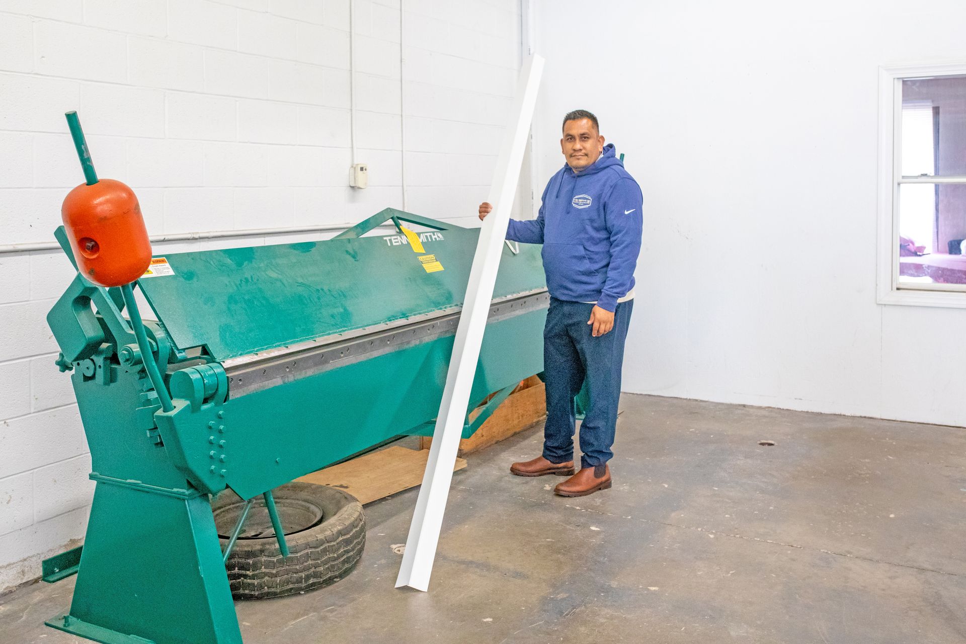 A person in a blue hoodie stands next to a large green metal brake machine holding a piece of white trim in a workshop.