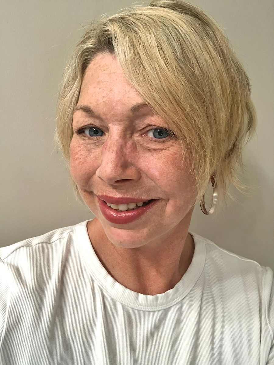 A person with short blonde hair and silver hoop earrings smiling at the camera in front of a neutral-colored wall.