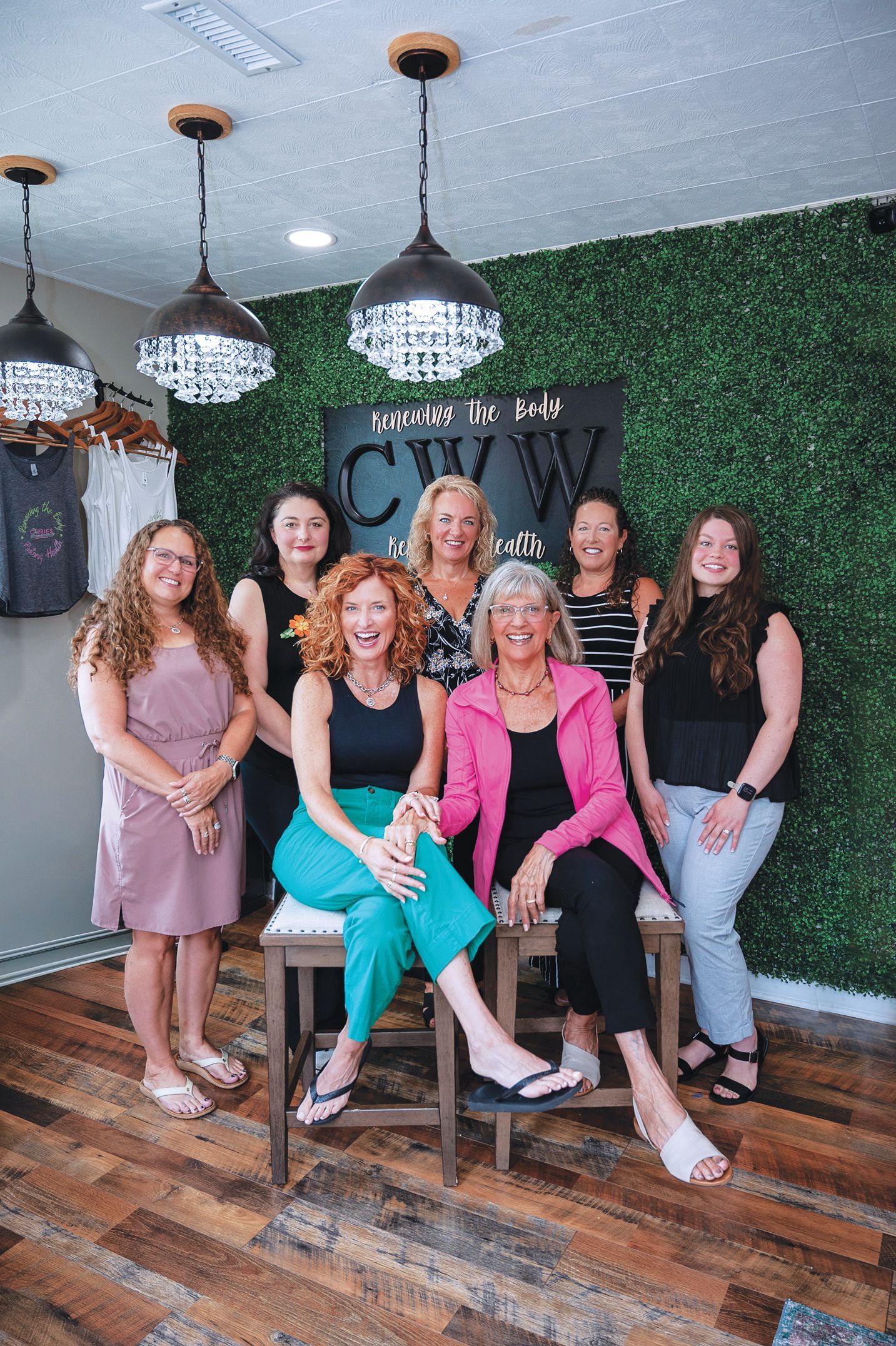 Seven women pose in front of a green decorative wall with hanging chandeliers in a clothing store setting.