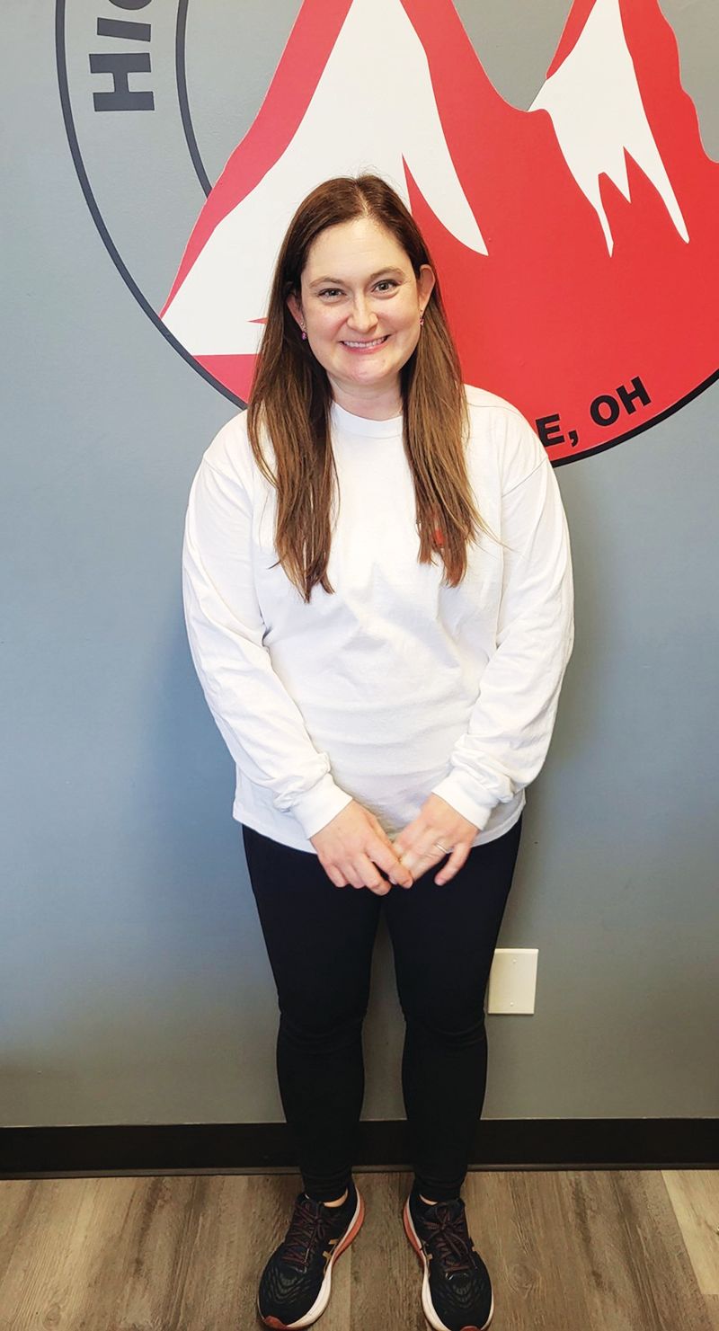 A person stands smiling in front of a gray wall with a logo featuring red mountains and the text 