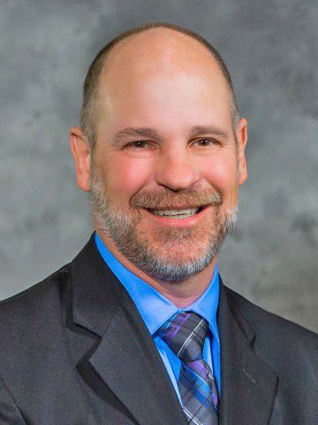 A smiling person wearing a dark suit, blue collared shirt, and a plaid tie against a gray, textured background.