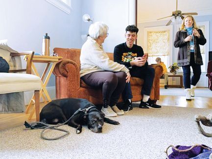 Two people talk on a couch while a black dog rests on the floor nearby, with another person standing in the background.
