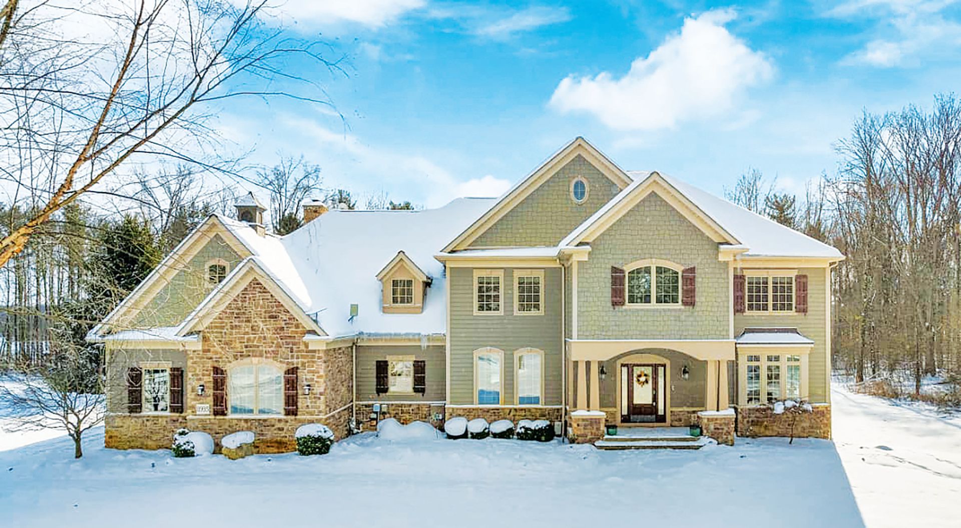 A two-story house with green siding and stone accents sits covered in snow against a backdrop of bare trees.