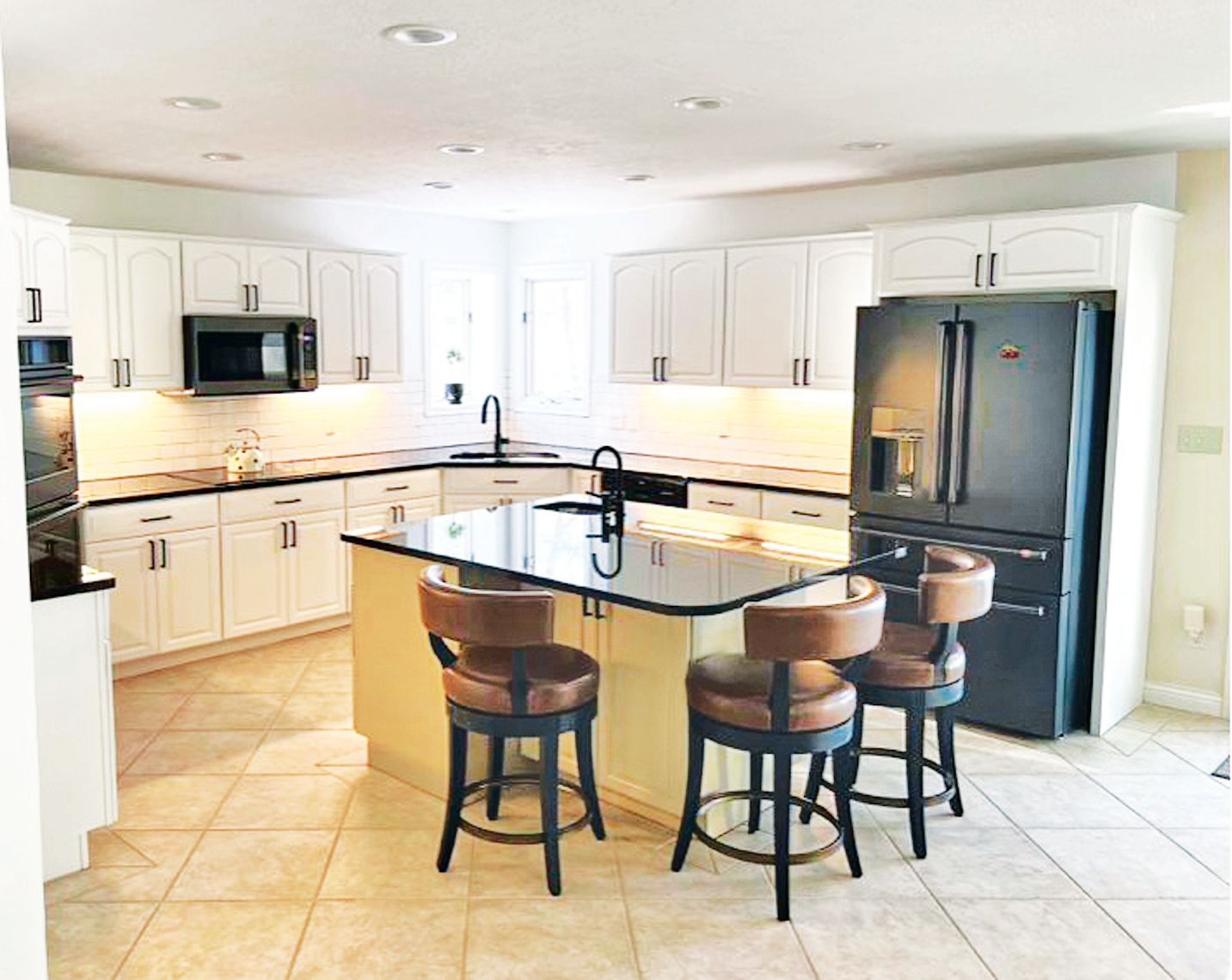 A modern kitchen with white cabinets, dark granite countertops, an island with three brown stools, and a black refrigerator.