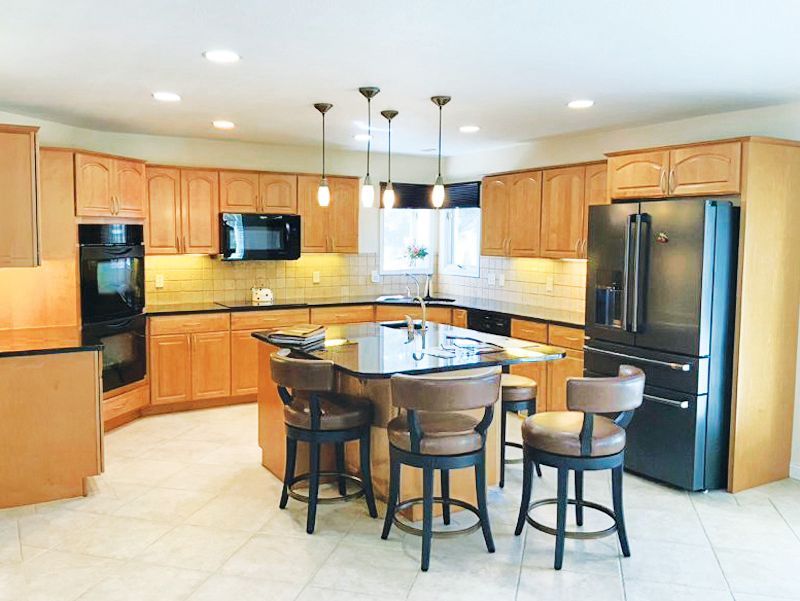 Modern kitchen with light wood cabinets, dark granite countertops, a center island with three stools, and black appliances.
