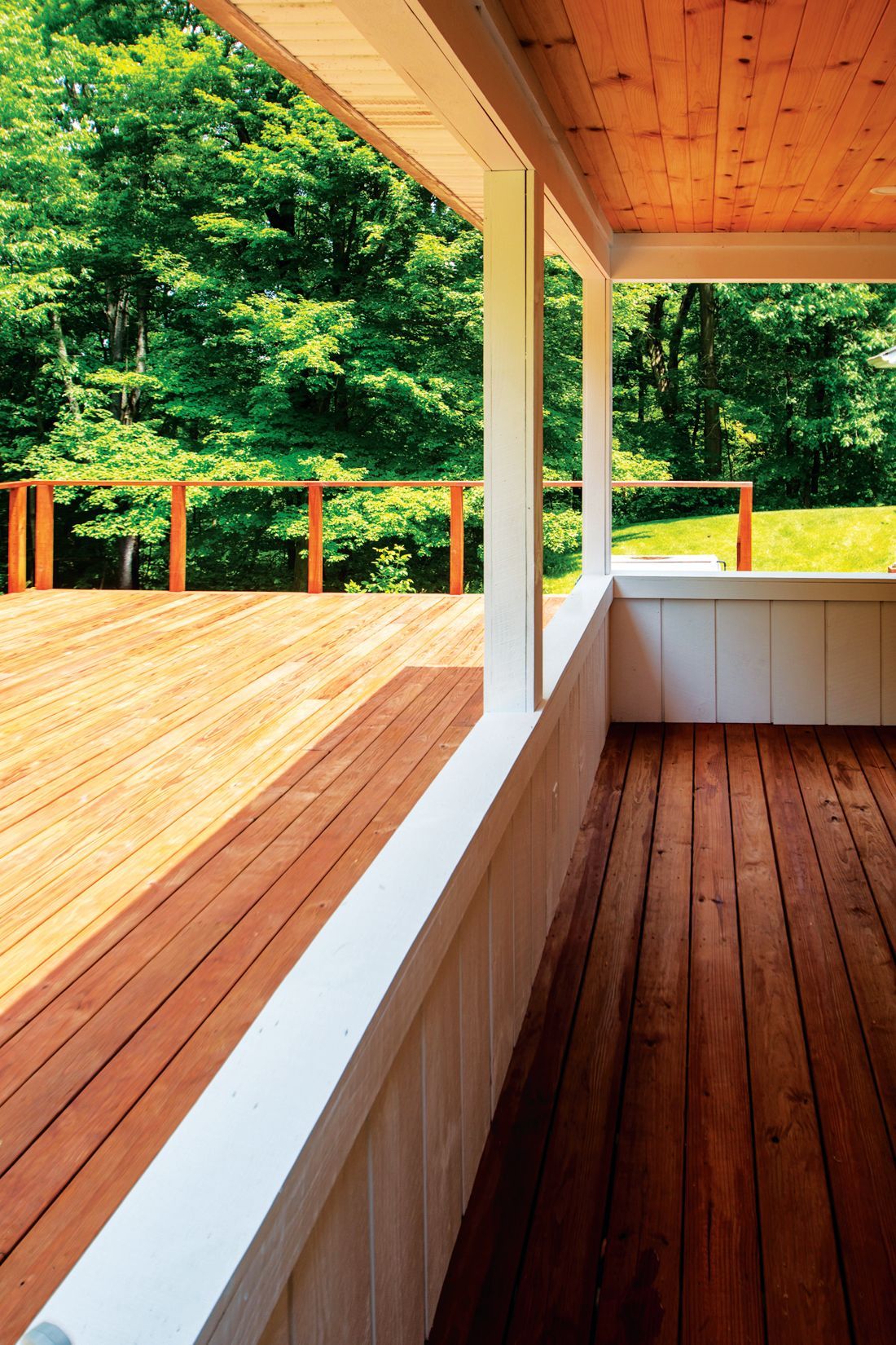 A covered wooden porch looking out onto a sunlit deck and lush green trees.