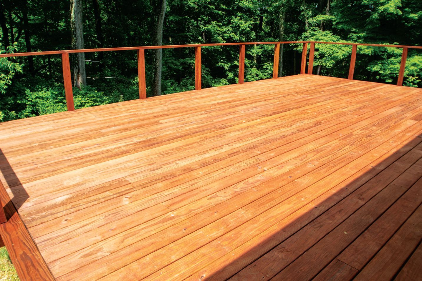 A wooden deck with a partially stained surface and a simple railing overlooking a dense green forest.