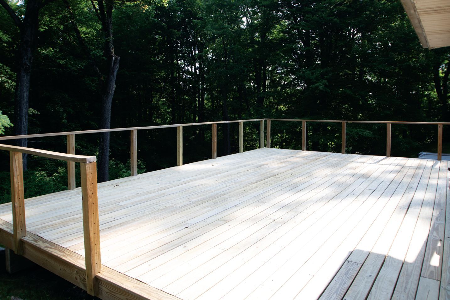 A wooden deck with railings extends outward against a backdrop of dense green trees.