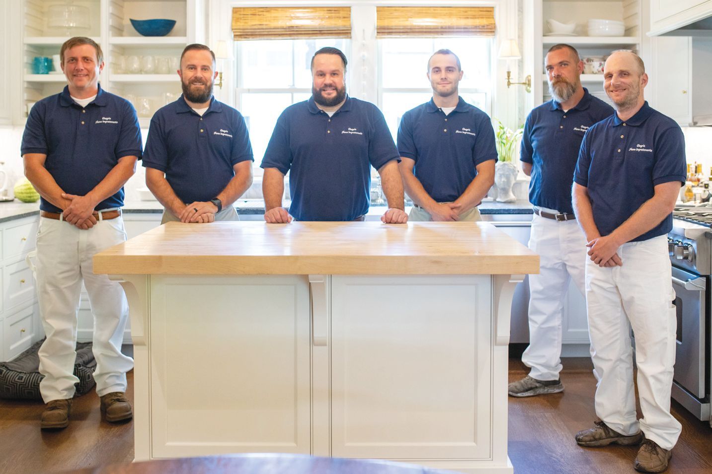 Six workers in matching navy blue polo shirts and white pants stand behind a wooden kitchen island in a residential kitchen.