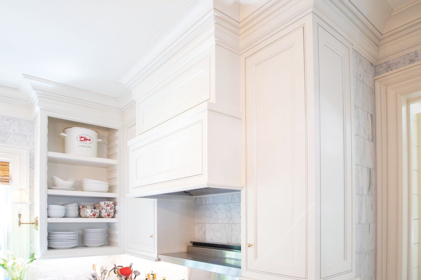 A white kitchen featuring a large custom range hood, tall cabinetry, and built-in shelving filled with white dishes.