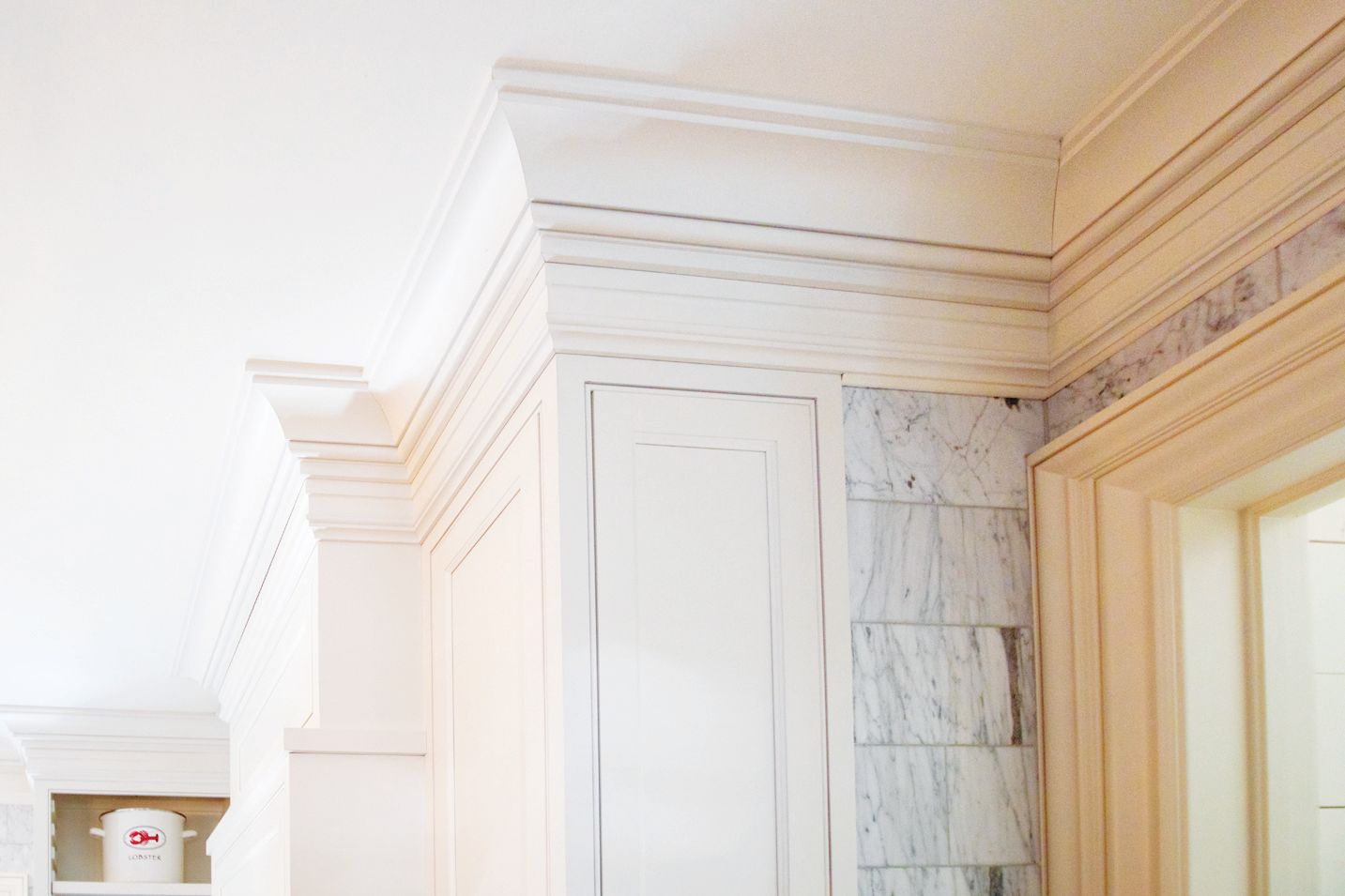 A corner of a white cabinet with decorative crown molding next to a marble-tiled wall and a doorway.