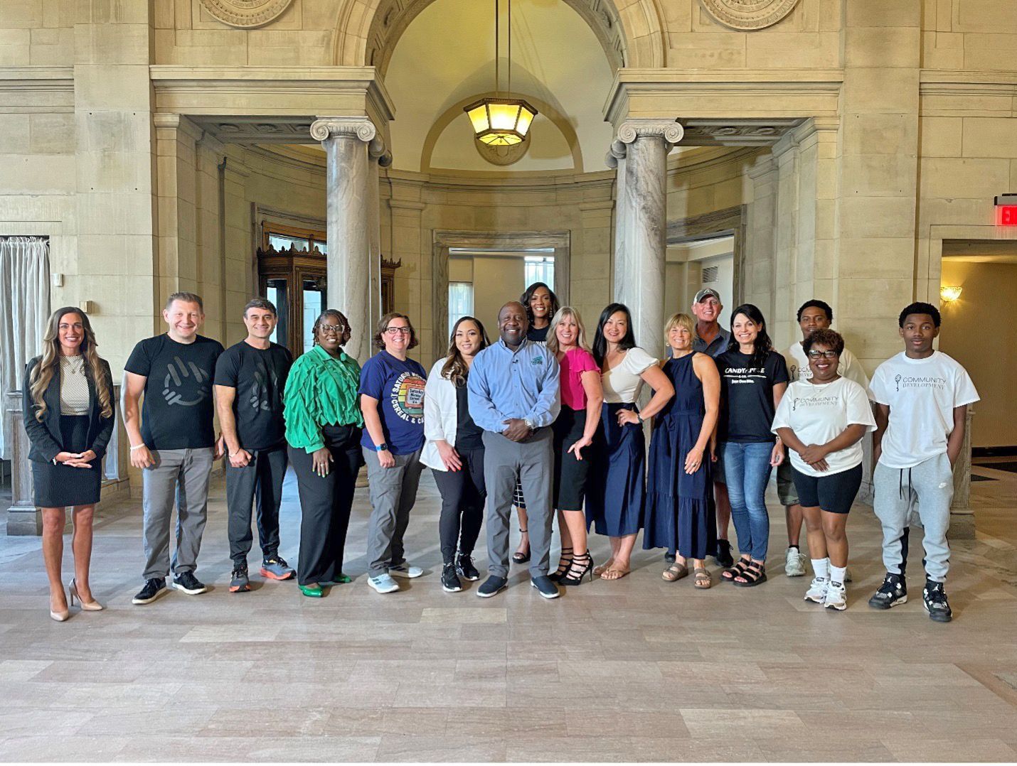A group of people standing in a grand, stone-arched hall with columns and a light fixture, smiling for a group photo.