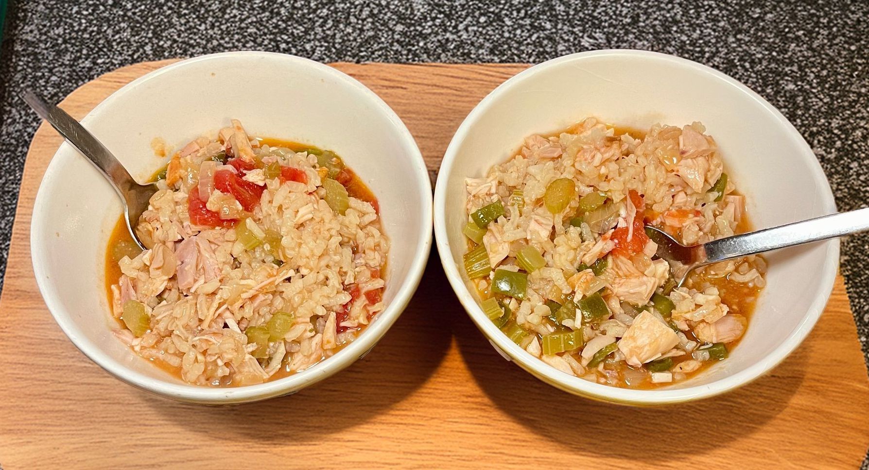 Two white bowls filled with chicken and rice stew, each with a metal spoon, resting on a wooden cutting board.