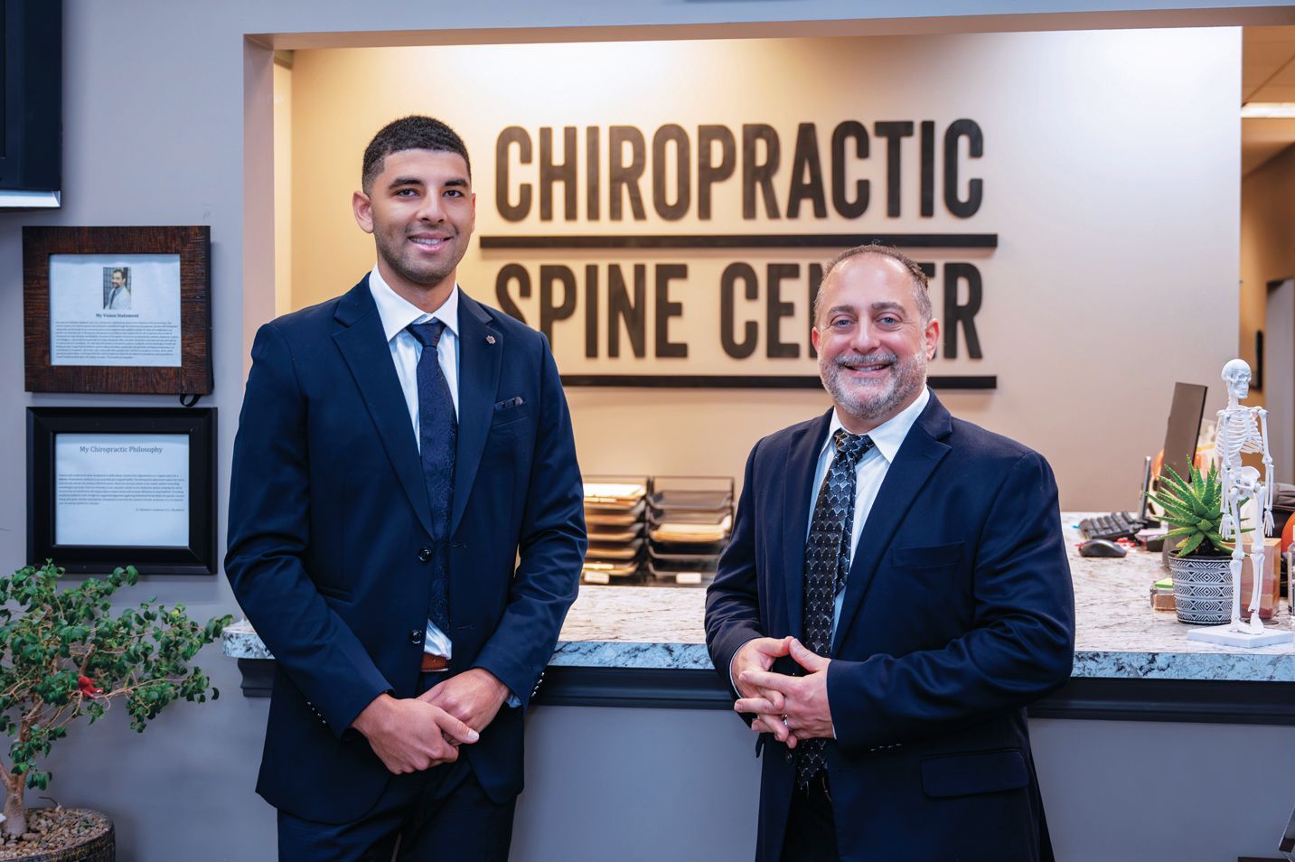 Two professionals in dark suits standing in front of a Chiropractic Spine Center sign inside an office.