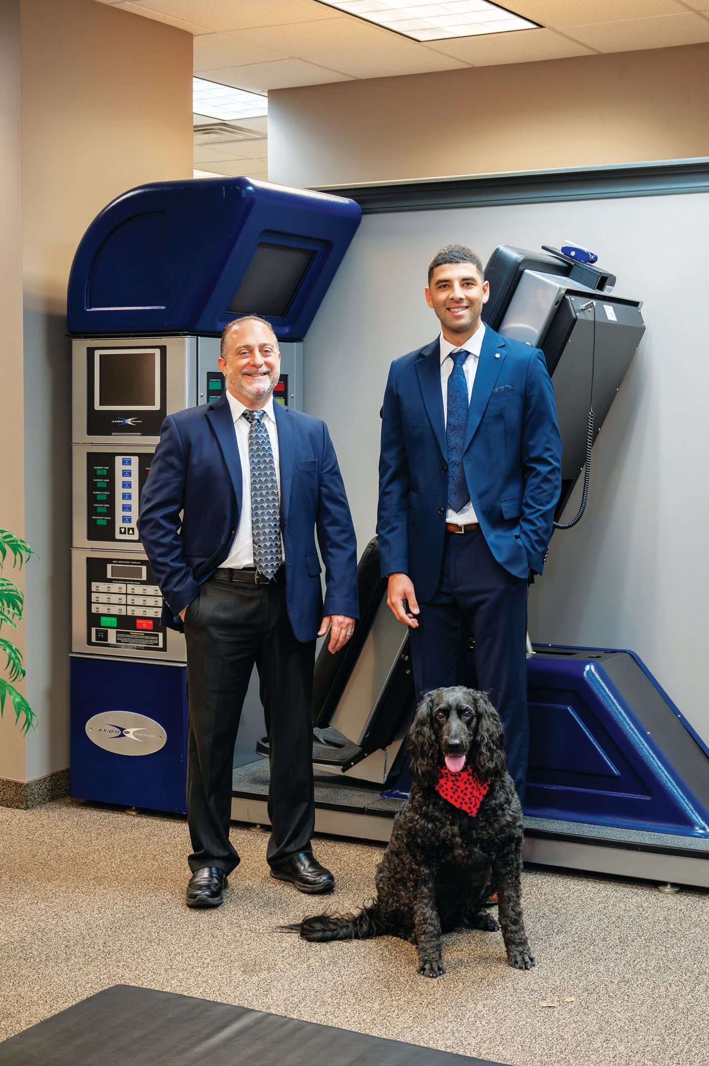Two men in suits stand next to a large blue medical device, with a black dog wearing a red bandana sitting in front.