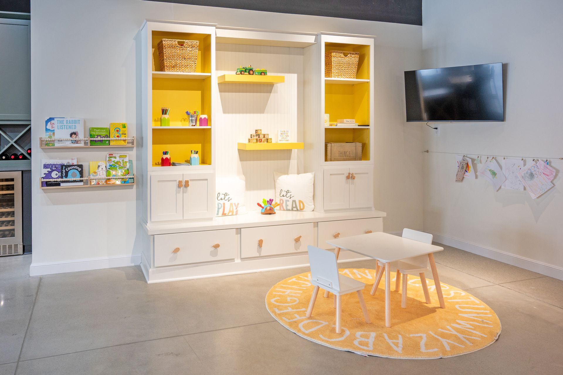 A white storage unit with yellow shelving, wall-mounted book ledges, and a small table and chairs on a yellow round rug.