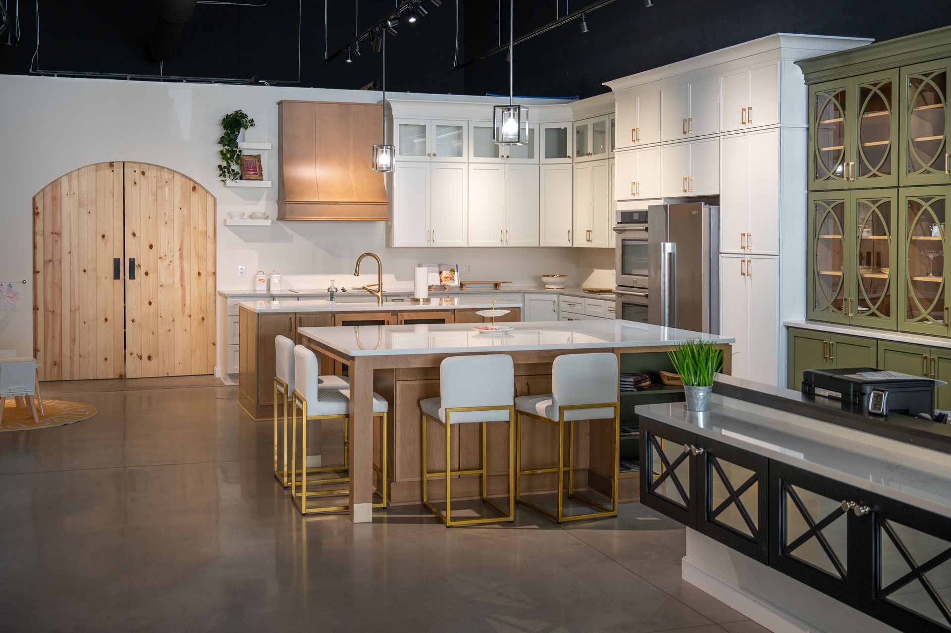 A kitchen showroom featuring a large central island, white cabinetry, a wood range hood, and green display cupboards.