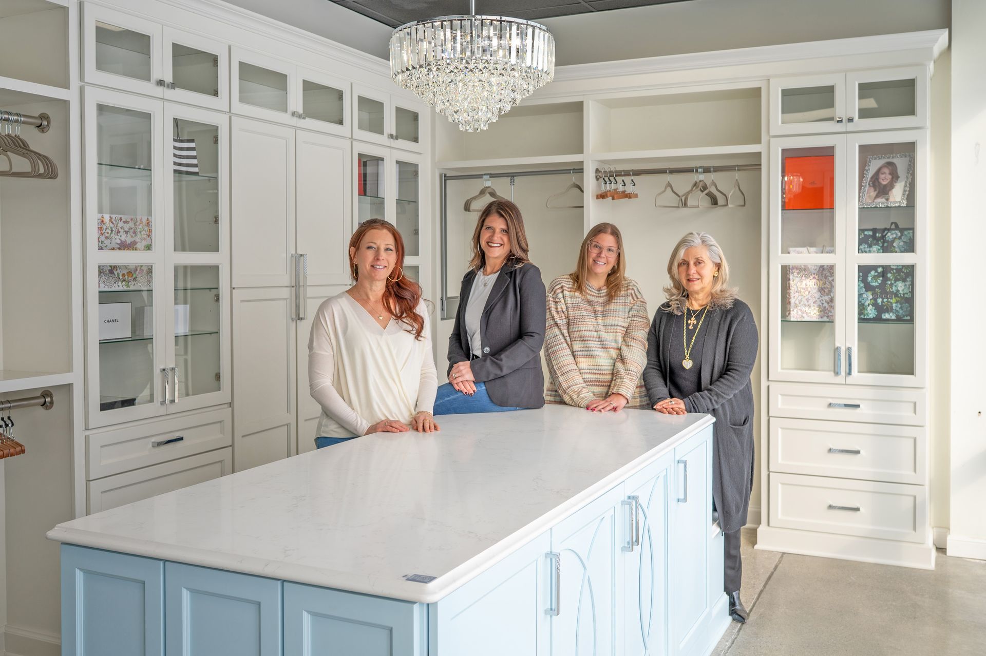Four people stand behind a light blue kitchen island in a white walk-in closet with a crystal chandelier.