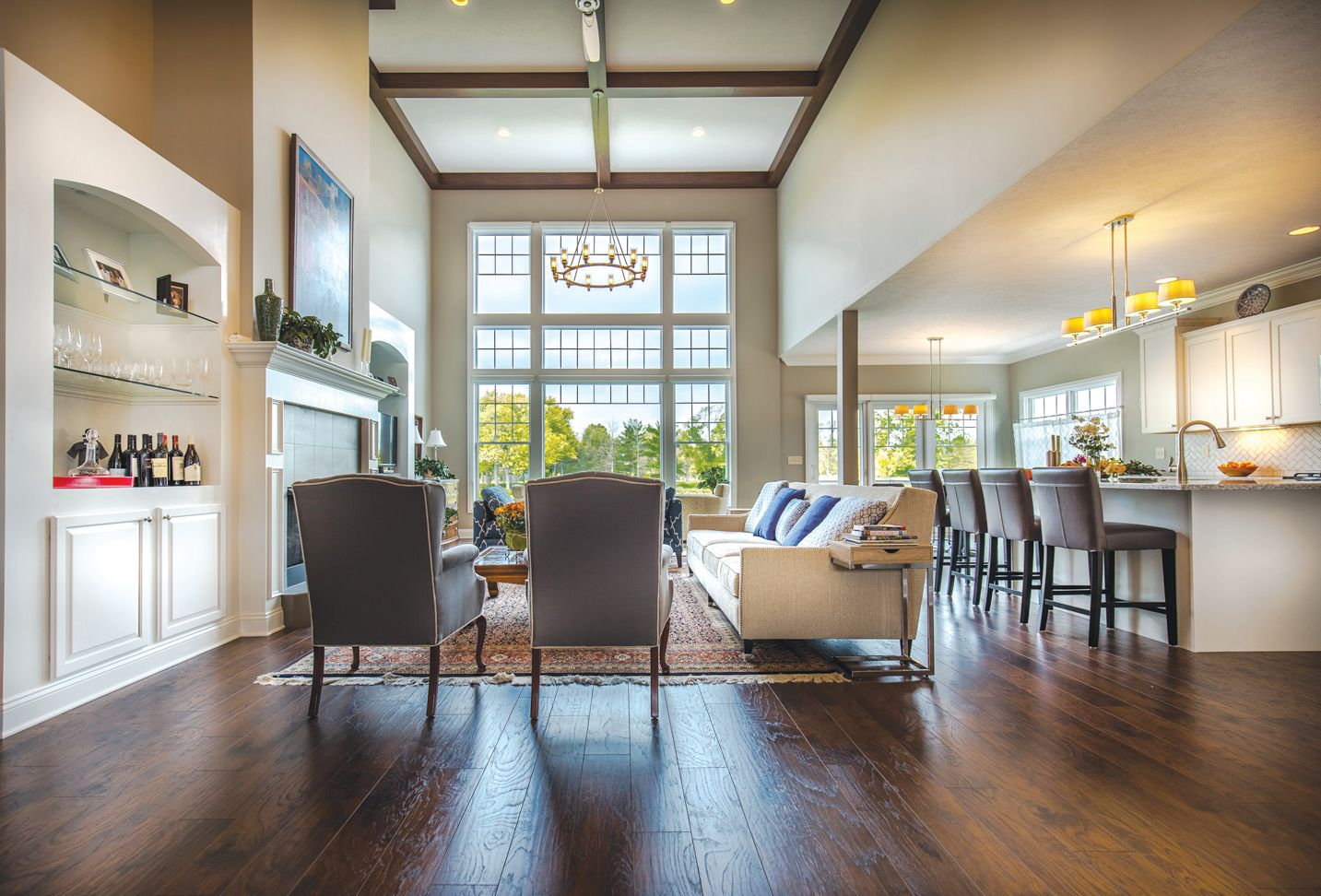 A bright, open-concept living area with dark wood floors, large windows, two gray chairs, a sofa, and a kitchen island.