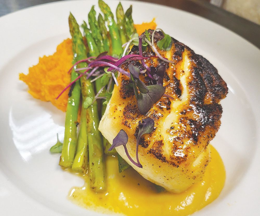 A seared white fish fillet with asparagus, served over orange vegetable mash and sauce, garnished with microgreens.