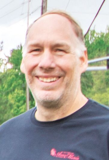 A smiling person wearing a dark blue t-shirt stands outdoors in front of a blurred green landscape.