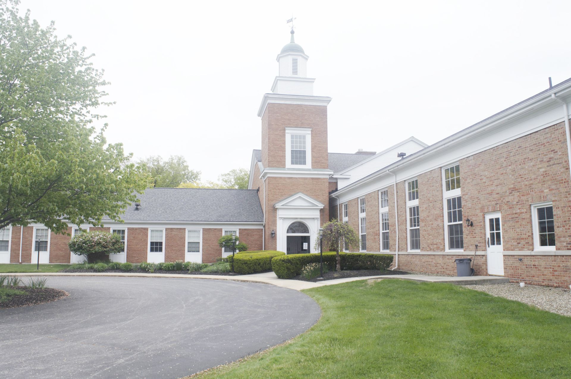 A brick building with a central bell tower and a white-trimmed entrance, set behind a circular driveway and lawn.