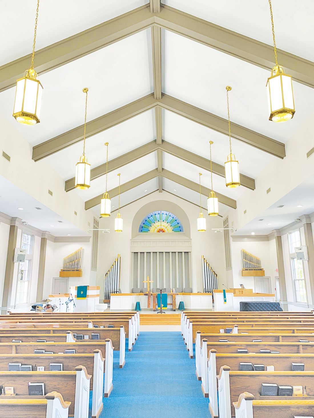 View of a church interior with wooden pews facing an altar, a blue carpeted aisle, and hanging lanterns under vaulted beams.