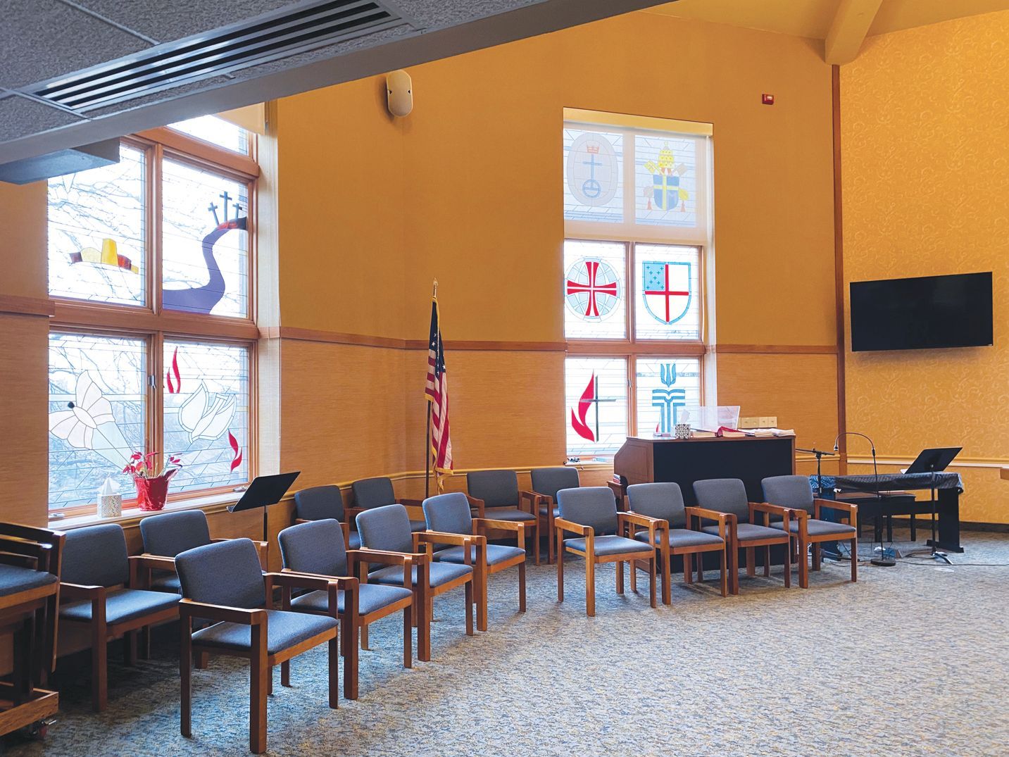 An interior view of a meeting space with a row of chairs facing a platform, American flag, and windows with stained glass.
