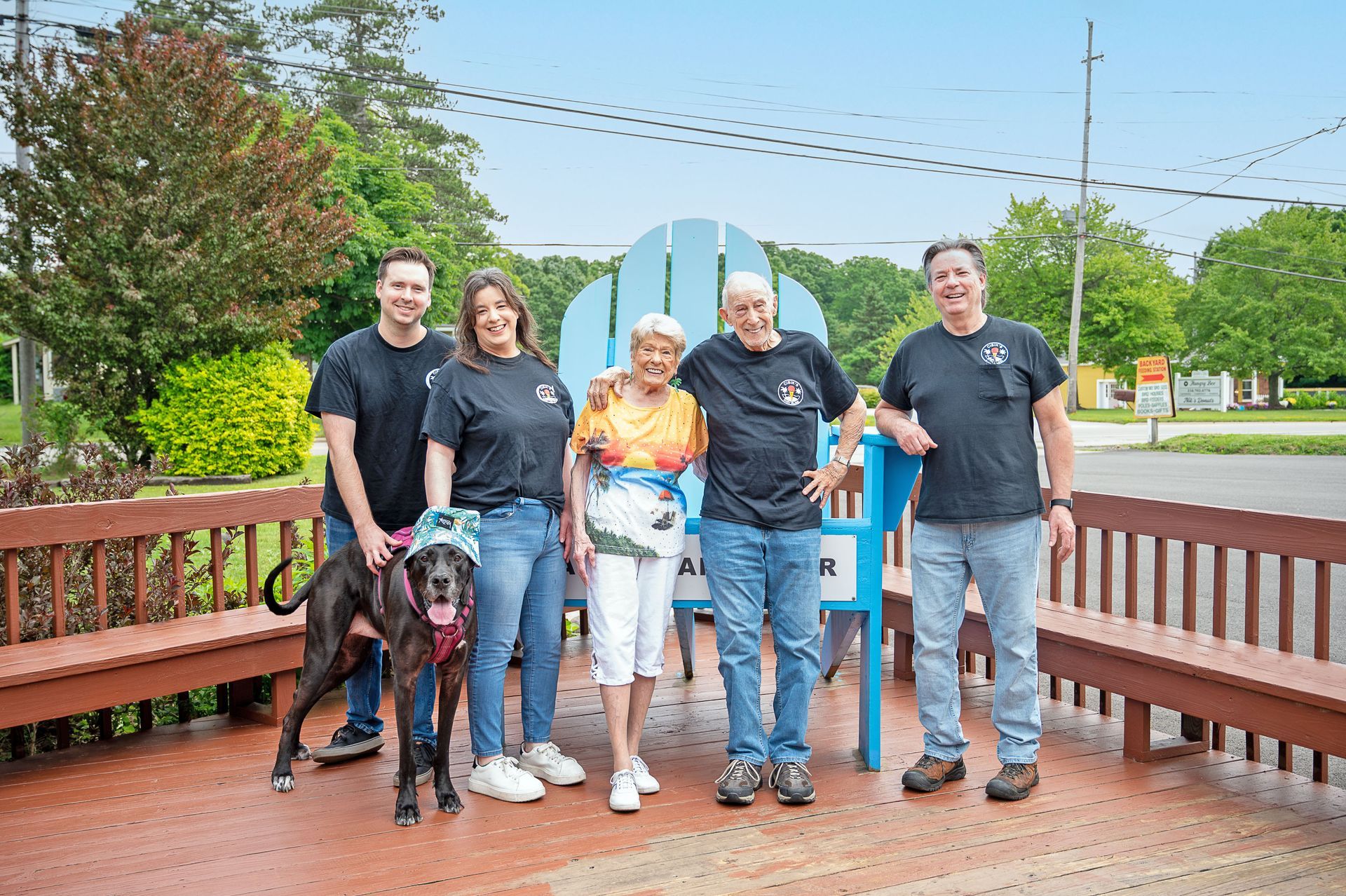 Five people and a dog stand on a deck in front of a large, blue wooden Adirondack chair in a bright outdoor setting.