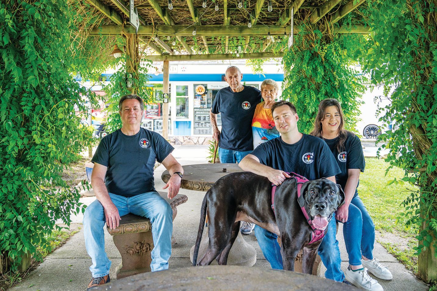 Four people and a large black dog sit beneath a vine-covered pergola on stone benches at a garden cafe.