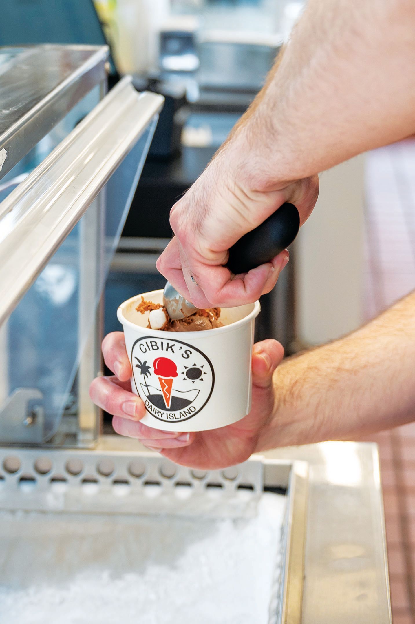 A person uses an ice cream scoop to serve a portion of ice cream into a small paper cup with a logo.