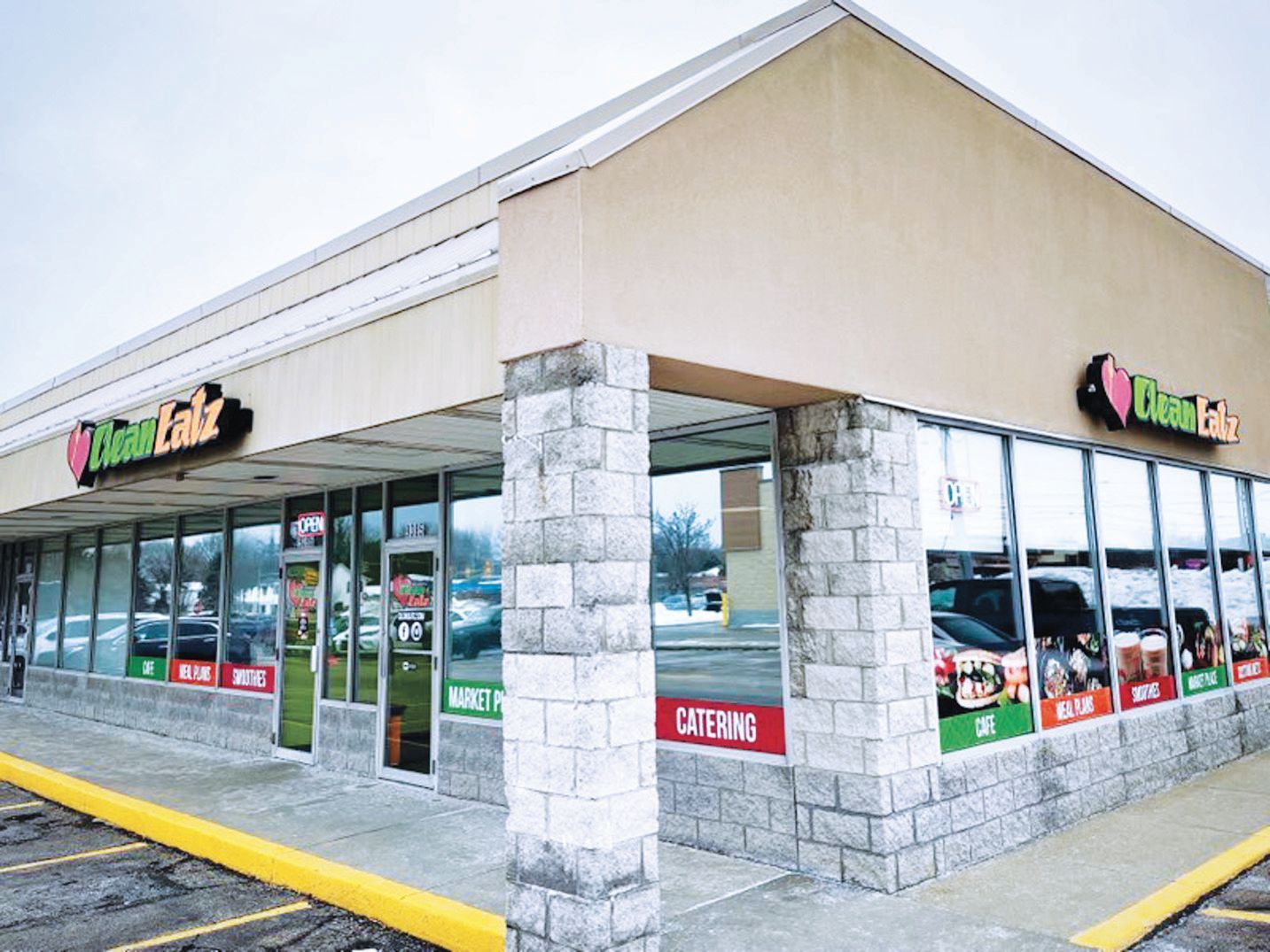 A Clean Eatz storefront with stone pillars, large windows, and branded signage in a shopping center parking lot.