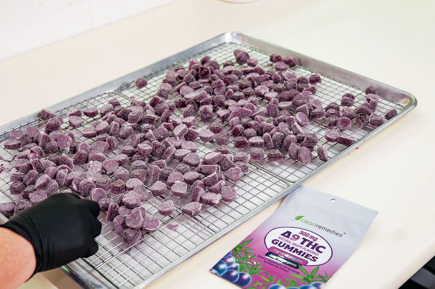 A gloved hand adjusts purple sugar-coated gummies on a wire drying rack next to a branded product pouch.