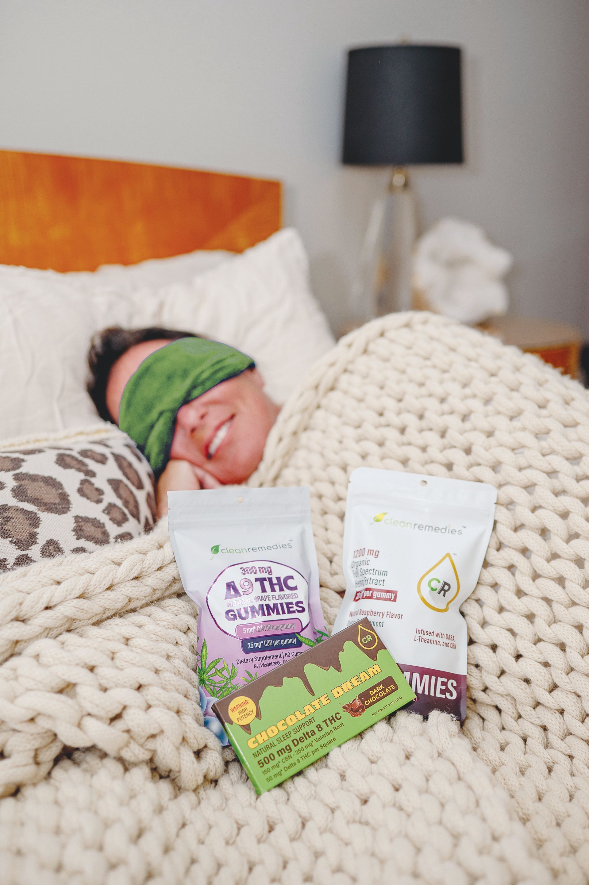 A person wearing a green eye mask rests in bed under a thick cream blanket with cannabis-infused snack packages in front.