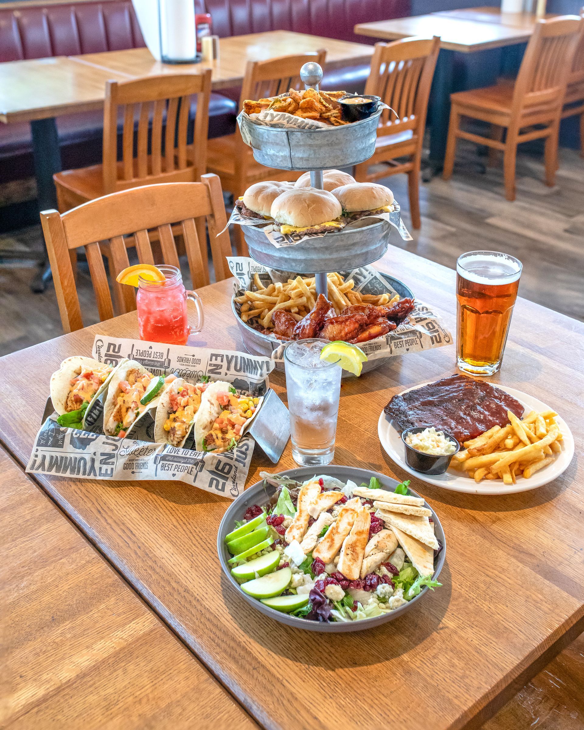 A wooden restaurant table spread with a tiered food stand, tacos, salad, ribs with fries, a cocktail, and a glass of beer.