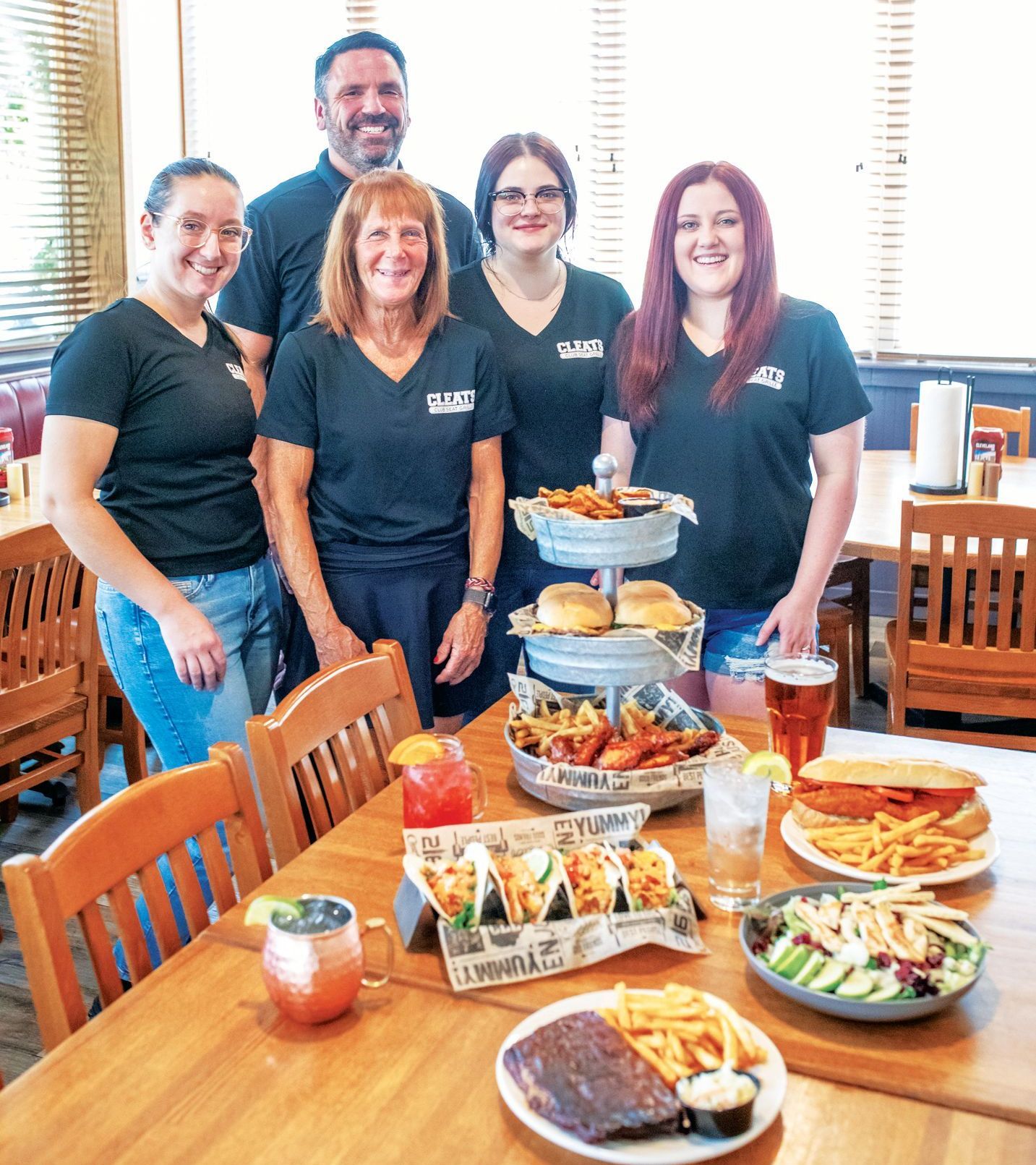Five people in black shirts stand behind a table laden with food and drinks in a restaurant setting.