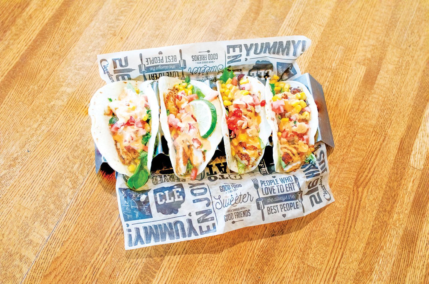 Four tacos filled with meat, cheese, and vegetables, served in a branded paper liner on a light wooden table.