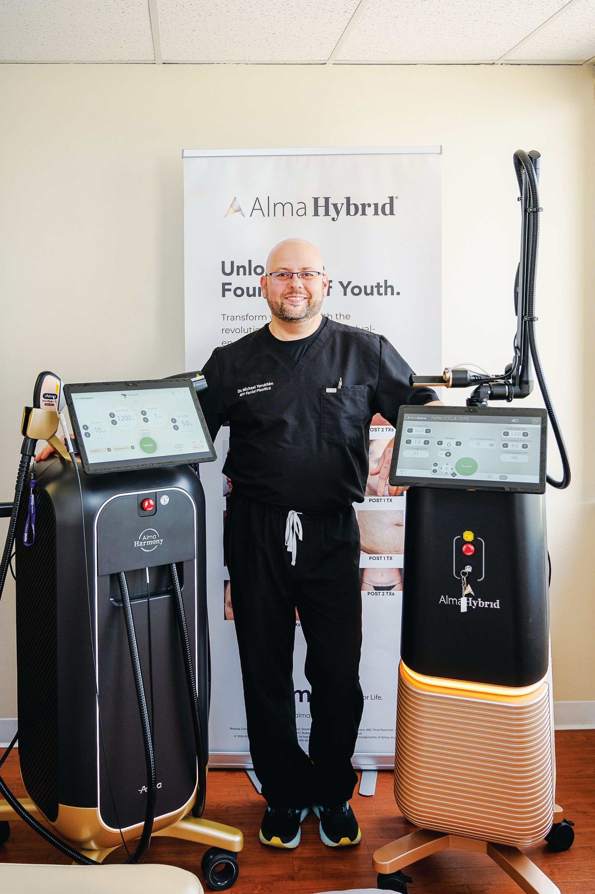 A professional stands smiling between two Alma medical aesthetic laser devices in a clinic setting.