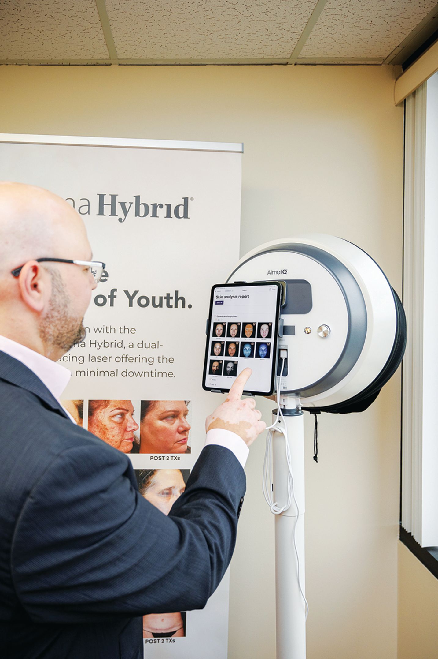 A man in a suit touches the screen of a medical imaging device for skin analysis next to a promotional display.