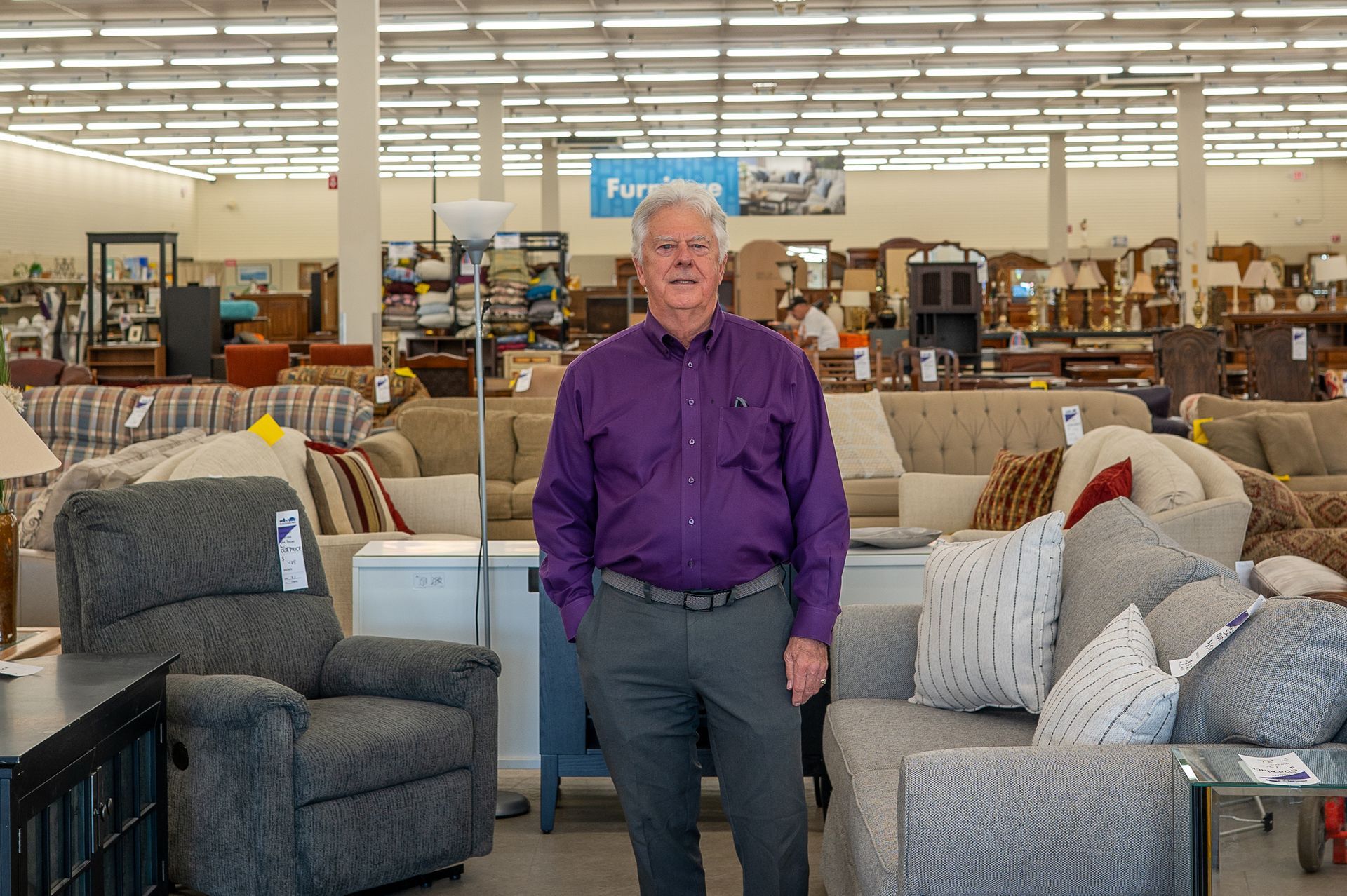 A person in a purple shirt and gray slacks stands in a furniture store, centered between a recliner and a sofa.