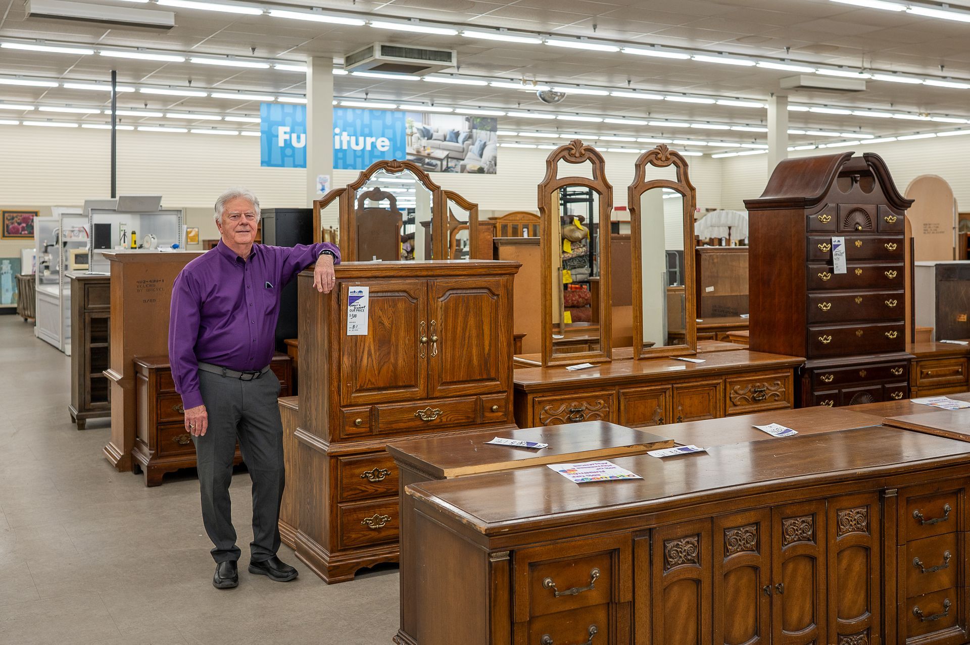 A person wearing a purple shirt and gray pants stands next to a wooden dresser in a furniture store.
