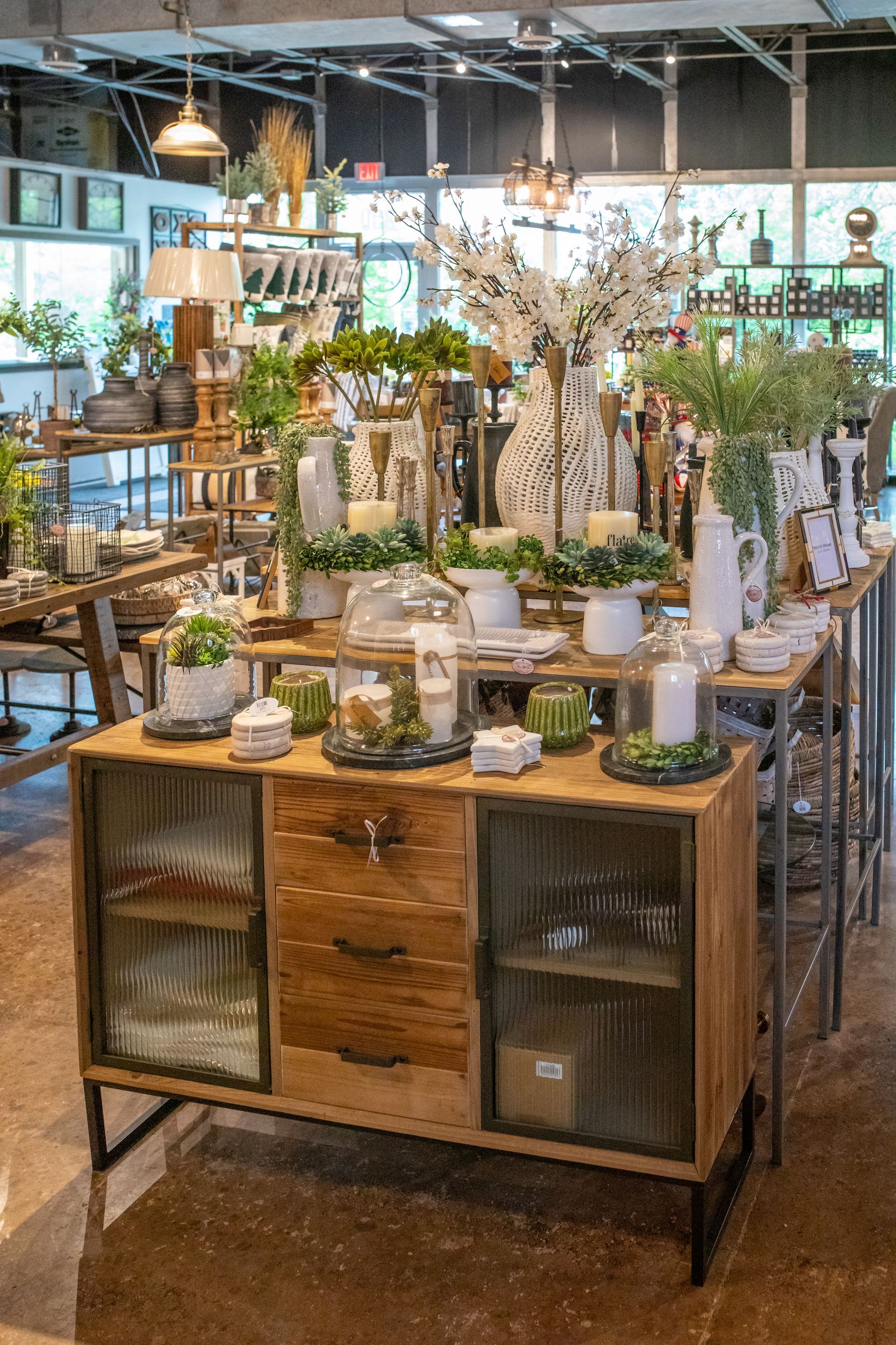 A rustic wooden sideboard with glass cabinet doors and drawers, topped with decorative plants and candles in a shop.