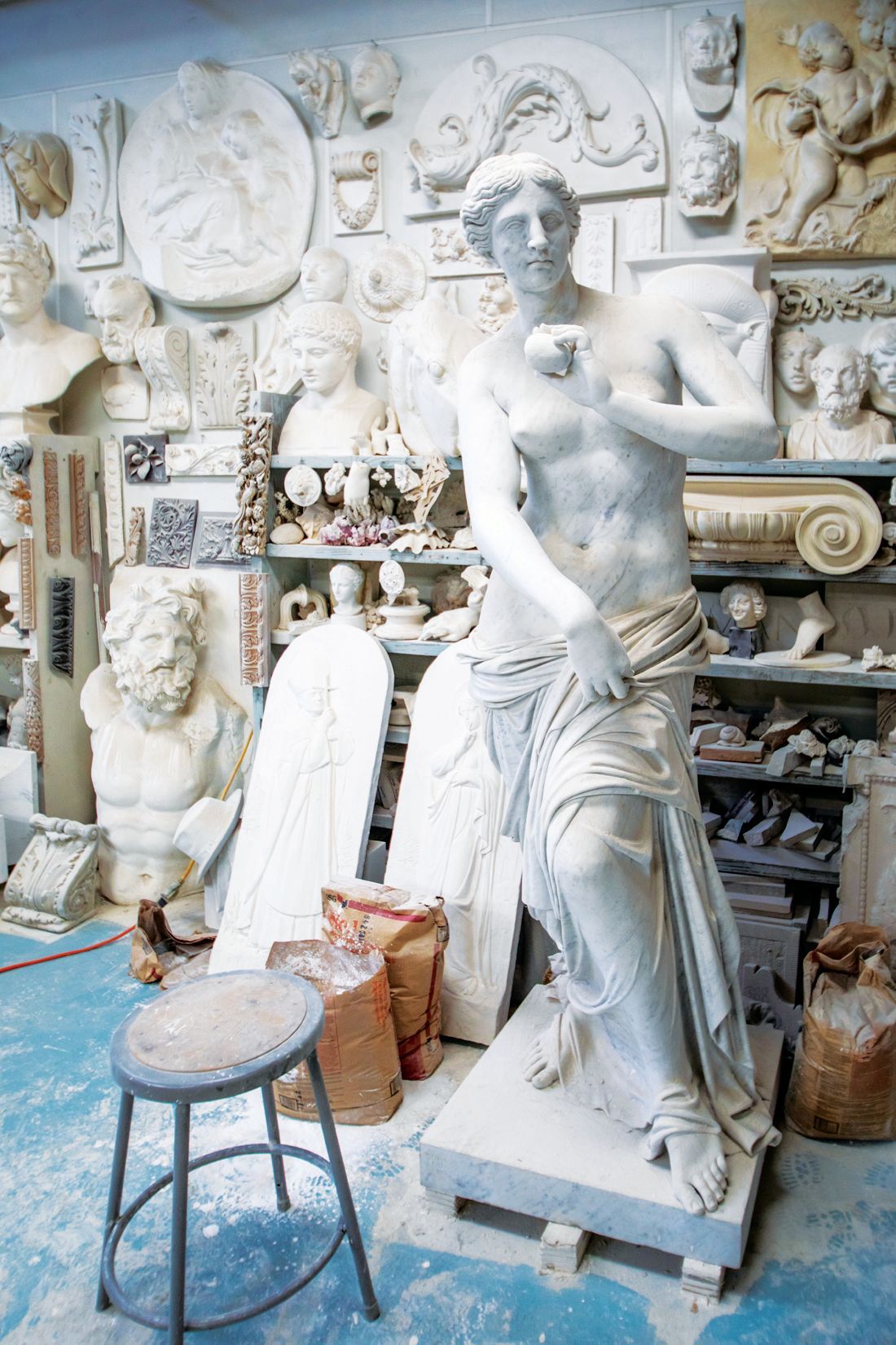 A plaster sculpture of a classical figure stands in a studio filled with various sculpted reliefs and busts.
