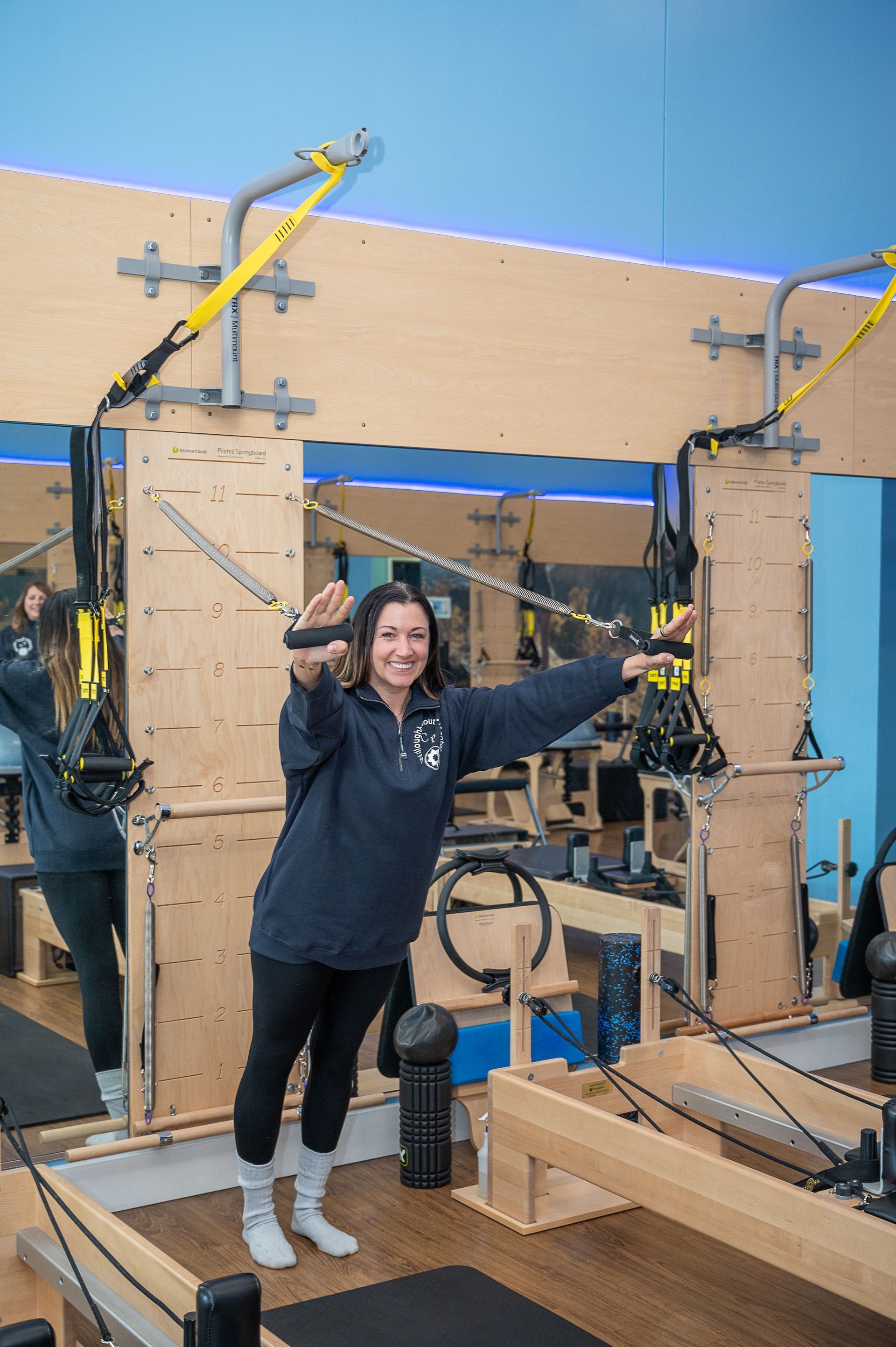 A smiling individual in a dark sweatshirt performing a suspension exercise in a light-wood Pilates studio.