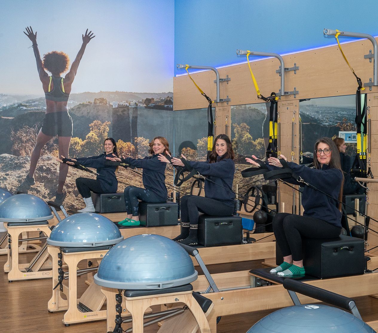 Four people sit on Pilates reformers in a gym, each holding onto TRX suspension straps with arms extended forward.