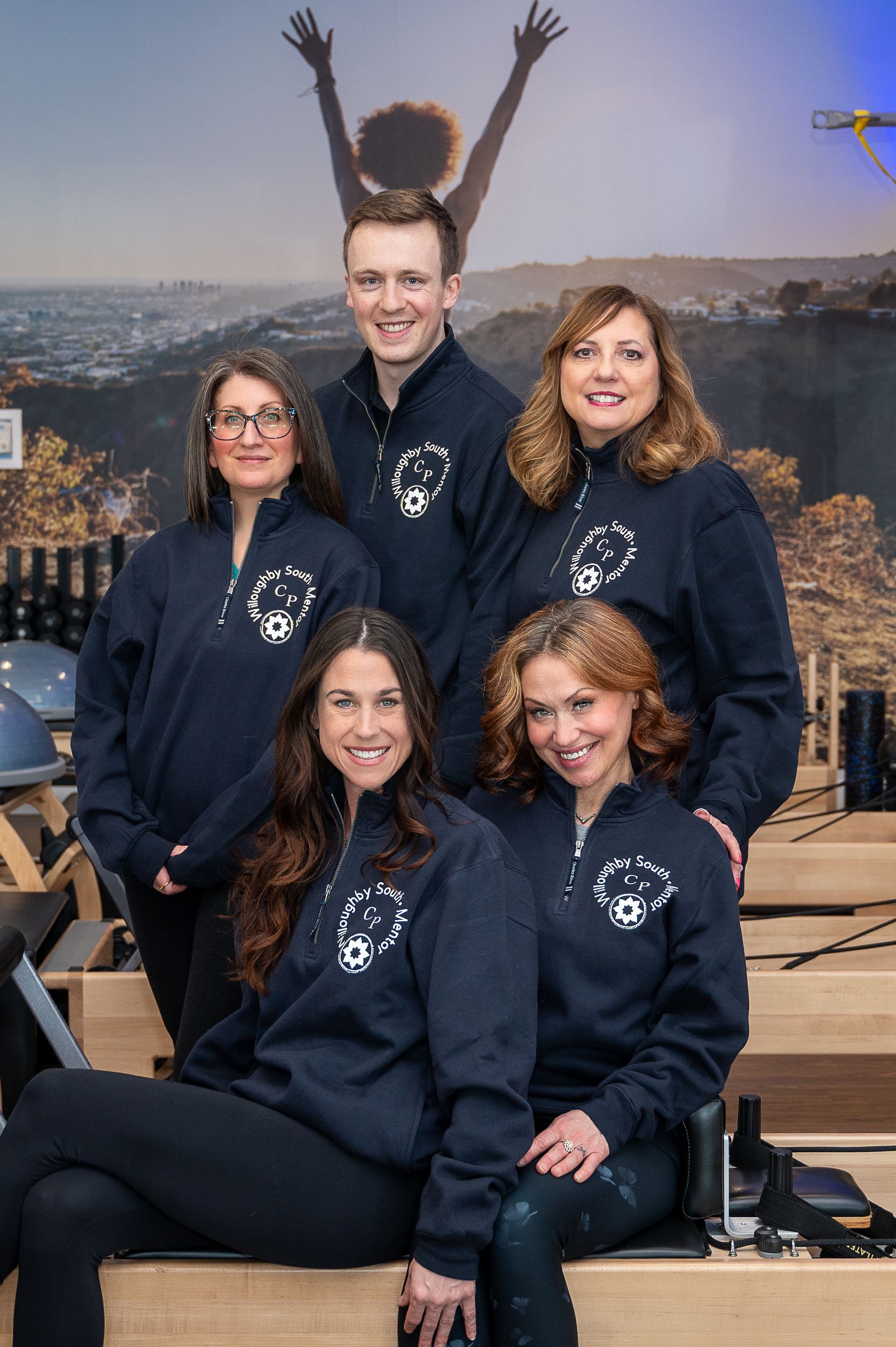 A group of five people wearing matching dark blue sweatshirts pose for a photo in a gym setting with a mural background.