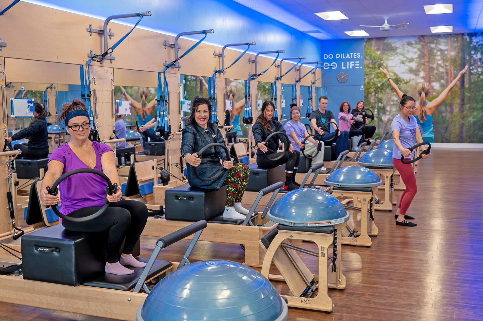 A group of people exercising on Pilates reformers and BOSU balls in a bright studio with a forest mural on the wall.